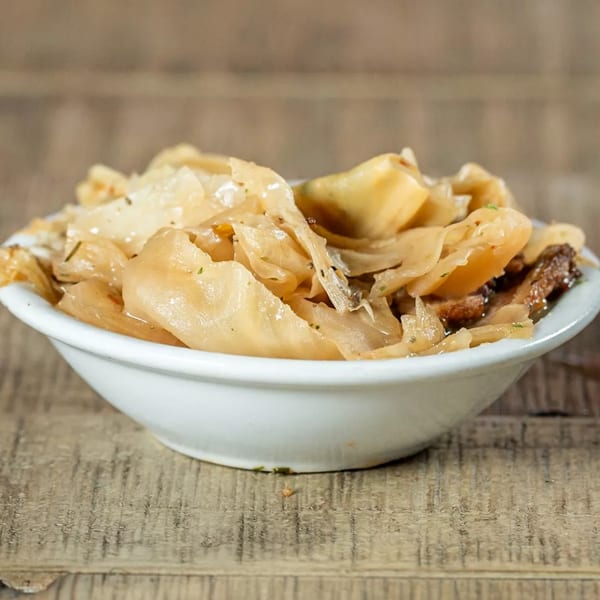 A white bowl of J's Eatery's soul-food fried cabbage, seasoned with pepper, sits on a wooden table.