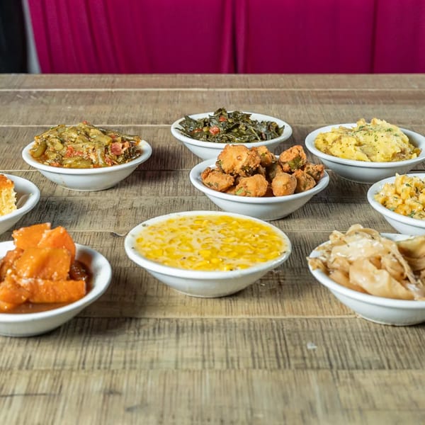 A wooden table laden with various soul food side dishes in white bowls, including greens, corn, and sweet potatoes.