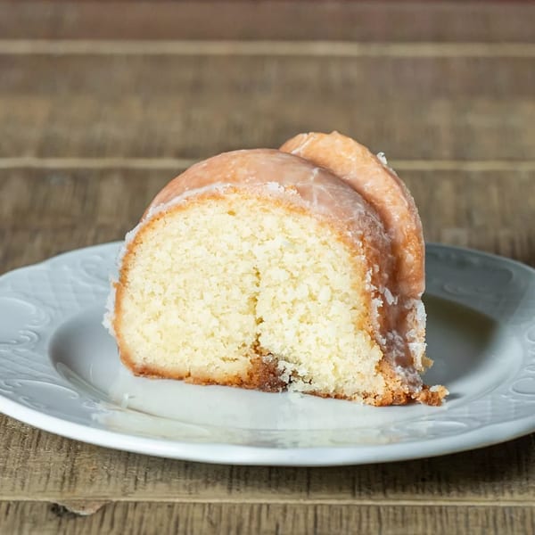 A slice of J's Eatery's glazed pound cake on a white plate, against a wooden background.